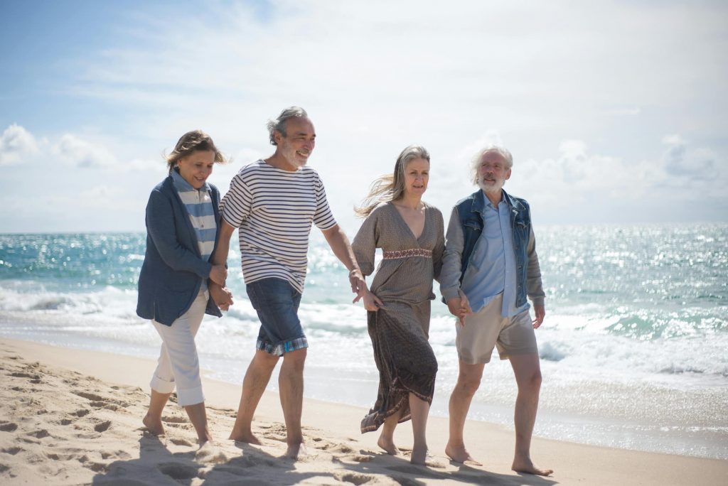 Vier ältere Erwachsene spazieren barfuß am Strand entlang, Hand in Hand, bei sonnigem Wetter und blauem Himmel.
